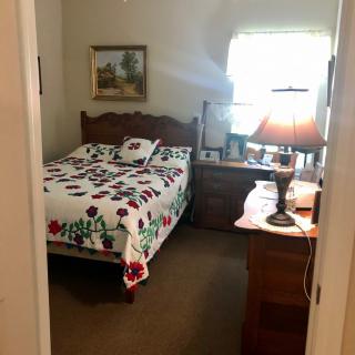 A cozy bedroom with a quilted bed and a nightstand topped with a lamp, under a softly lit window.
