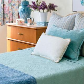 A cozy bedroom scene with a neatly made bed, decorative pillows, and a wooden dresser adorned with a vase and flowers.