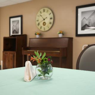 A cozy dining area featuring a simple table setting with flowers, a wall clock, and a piano in the background.
