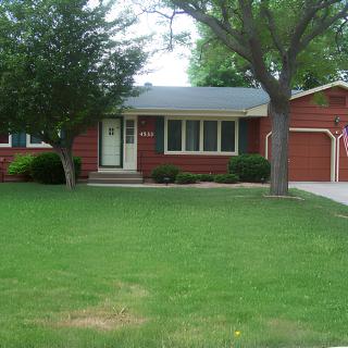A red brick suburban house with a well-kept lawn and a large tree.