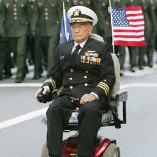 A military veteran in formal uniform participates in a parade while seated in a motorized wheelchair, holding an American flag.