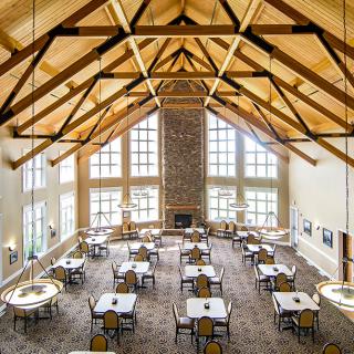 A spacious dining hall with wooden beams and large windows, featuring neatly arranged tables and chairs.