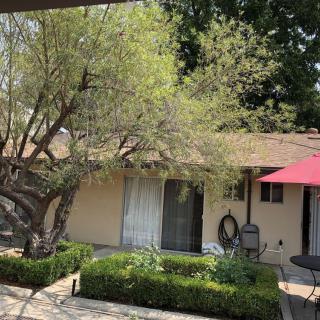 A cozy outdoor patio area with a tree and garden in front of a house, shaded by a red umbrella.