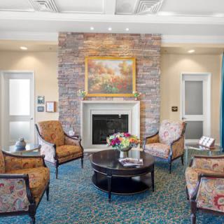 A cozy living room with a fireplace, surrounded by elegant chairs and a central coffee table adorned with a floral arrangement. The walls feature a serene painting above the mantel.