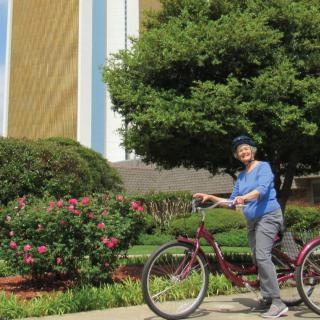 A person standing with a bicycle in a garden setting with trees and a building in the background.