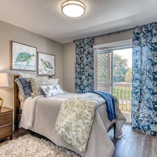 A cozy bedroom with blue and beige decor, featuring a single bed, artwork on the walls, and a window with floral curtains.