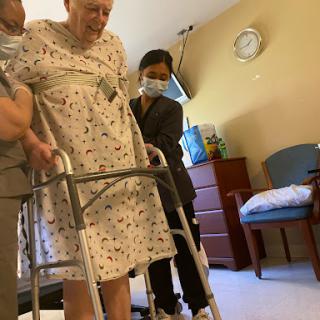 Two caregivers assist an elderly person using a walker in a healthcare setting.