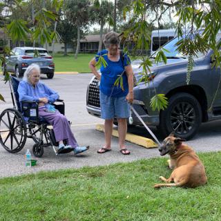 A caregiver takes a moment with a senior citizen in a wheelchair while their dog enjoys some time on the grass.