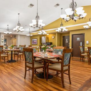 A warmly lit dining area with wooden floors, elegant chandeliers, and neatly arranged tables and chairs.