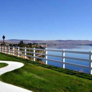 A scenic view of a lake with a white fence and pathway in the foreground, surrounded by green grass and distant hills under a clear blue sky.