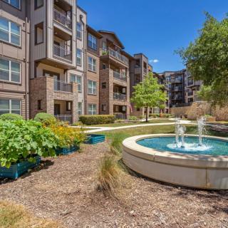 A modern apartment complex with a landscaped garden and fountain under a clear blue sky.