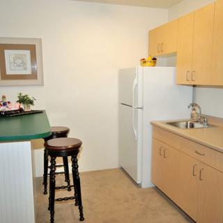 A compact apartment kitchen with wooden cabinets, a white fridge, and bar stools at the counter.