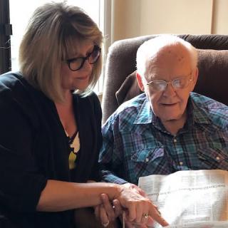 A woman and an elderly man reading a book together indoors.