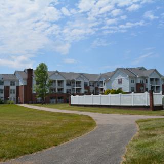 A modern apartment complex with a pathway and landscaped surroundings under a cloudy sky.