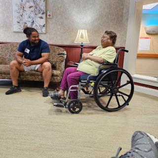 An elderly woman in a wheelchair engages in conversation with a caregiver in a waiting area.