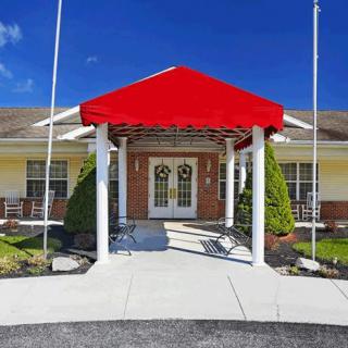 A building entrance with a red awning, supported by white pillars and surrounded by well-maintained landscaping.