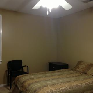 A minimalist bedroom with a bed, nightstand, and chair under a ceiling fan.
