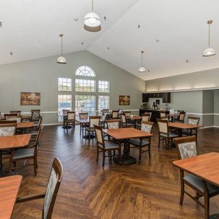 A spacious dining hall with multiple tables and chairs arranged on a wooden floor, illuminated by overhead light fixtures.
