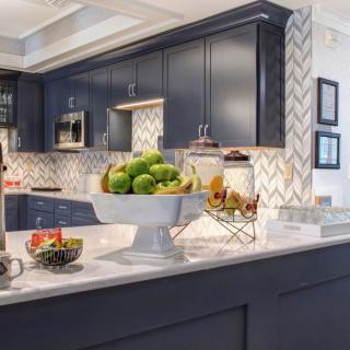 A modern kitchen with dark cabinets, a chevron backsplash, and a fruit display on the island.
