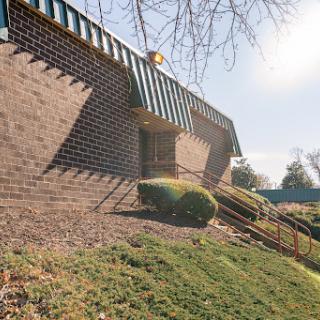 A side view of a brick building with sunlight casting shadows, featuring decorative overhangs and landscaped surroundings.