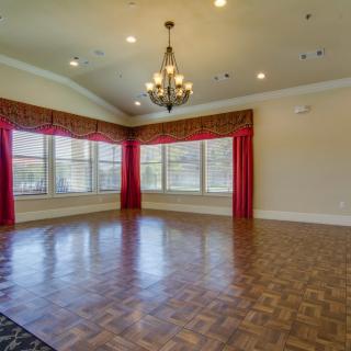 An empty room with hardwood flooring, large windows adorned with red curtains, and a chandelier hanging from the ceiling.