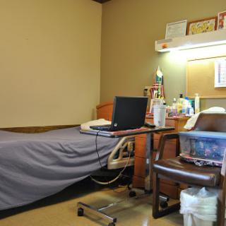 A view of a patient room in a hospital, featuring a bed, desk with a laptop, and various medical supplies.