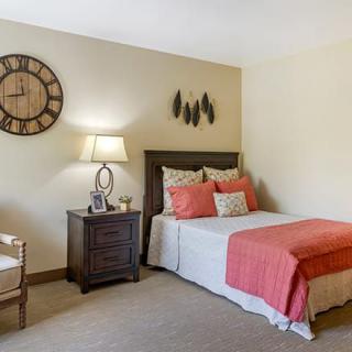A cozy bedroom with a neatly made bed, accentuated by a coral throw blanket and cushions. A wooden side table holds a lamp, and a large decorative clock adorns the wall.