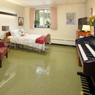 A cozy room featuring a piano, hospital bed, and a comfortable chair with sunlight streaming through the window.