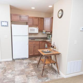 A small apartment kitchen with wooden cabinets, a refrigerator, and a microwave. A table and chair are set against the wall.