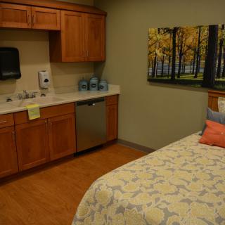 A cozy room with a bed and kitchenette, featuring wooden cabinets and a nature-themed wall art.