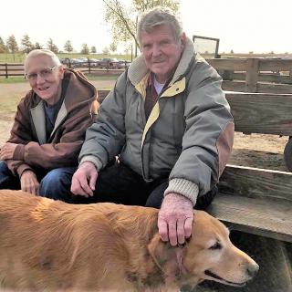 Two men sitting on a bench outdoors, petting a dog.