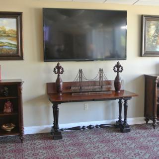 A living room setup featuring a large wall-mounted television, surrounded by framed artwork and decorative pieces on wooden furniture.