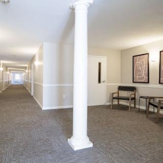 A well-lit interior hallway featuring a column, carpeted floor, and artwork on the walls.
