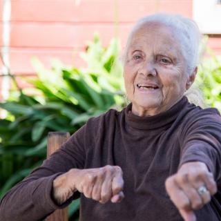 An elderly woman smiling while sitting outside surrounded by greenery.