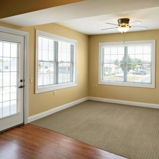 A bright empty room with a ceiling fan, large windows, and a mix of carpet and wood flooring.