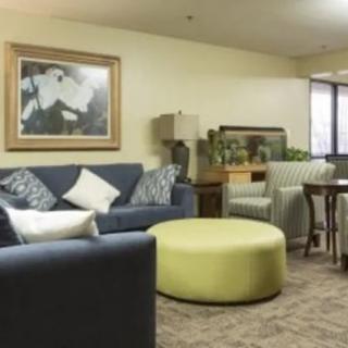 A cozy living room with a blue sofa, green ottoman, patterned armchair, and a framed painting on the wall.