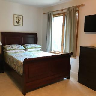 A cozy and well-furnished bedroom featuring a wooden bed, natural light from the window, and a mounted television.