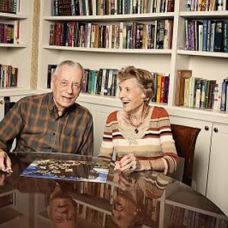 An elderly couple smiling while working on a puzzle at a table in front of a bookshelf.