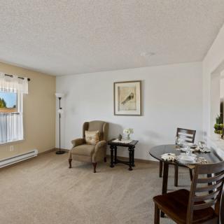 A cozy living room with a comfortable armchair, side table, dining table with chairs, and a large window for natural light.