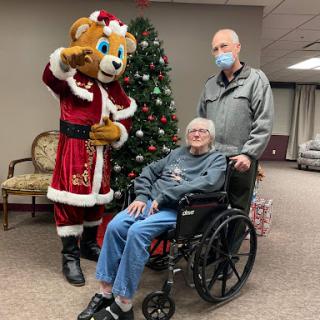 A festive moment captured with a cheerful mascot, an elderly individual in a wheelchair, and a companion, celebrating the holiday season.