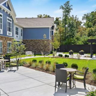 A sunny outdoor patio area with tables and chairs, surrounded by well-maintained landscaping and adjacent to a residential building.