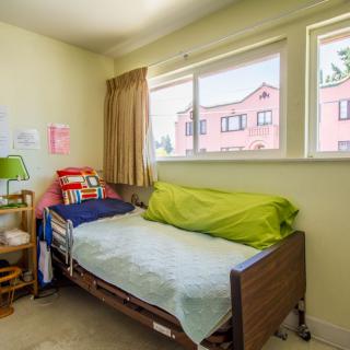 A cozy bedroom with a single bed by the window, featuring colorful bedding and a lamp on a small table.