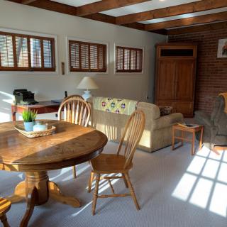 A cozy living room with a wooden dining table and chairs, a sofa, and ample natural light coming through the windows.