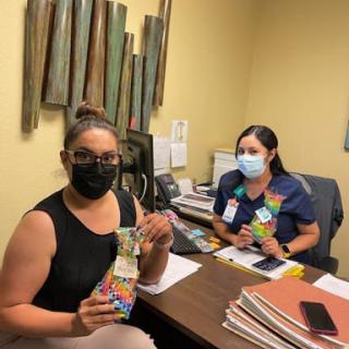 Two women in an office environment, both wearing masks and holding colorful items.