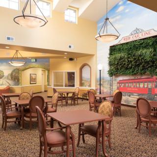 A spacious dining room with elegant tables and chairs, illuminated by chandeliers and natural light, featuring decorative wall art.