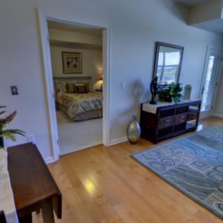 A cozy living room with hardwood floors, decorated with a large rug and stylish furniture, leading into a bedroom.