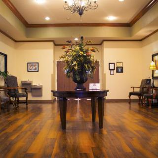 A warmly decorated lobby area with wooden flooring, a central table with floral arrangement, and seating along the walls.