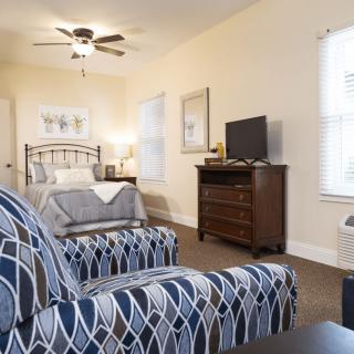 A cozy bedroom featuring a bed, patterned chair, dresser with a TV, and soft lighting.