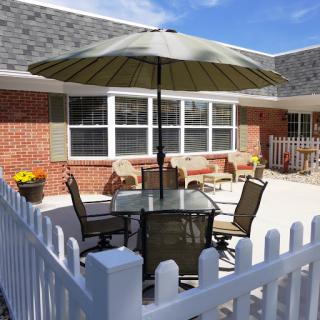 A cozy outdoor patio with a table and chairs beneath an umbrella, surrounded by a white picket fence.