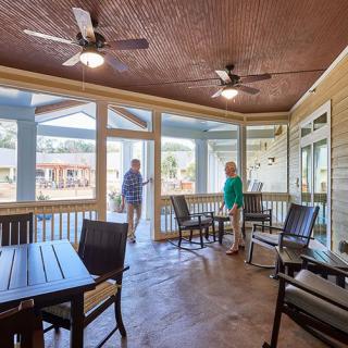 A screened porch with tables and chairs, featuring ceiling fans, overlooks a scenic outdoor area, with two people enjoying the view.